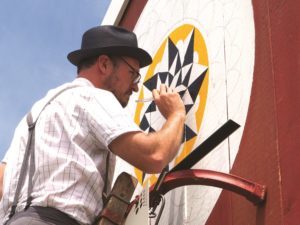 A man in short sleeve white shirt, suspenders and brown brimmed hat stands on a ladder as he paints a barn star or hex sign on a red barn.