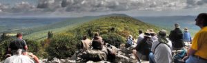 People sit on rocks with their backs to the camera, looking out over mountains as they watch for raptors during the fall hawk migration at Hawk Mountain Sanctuary.
